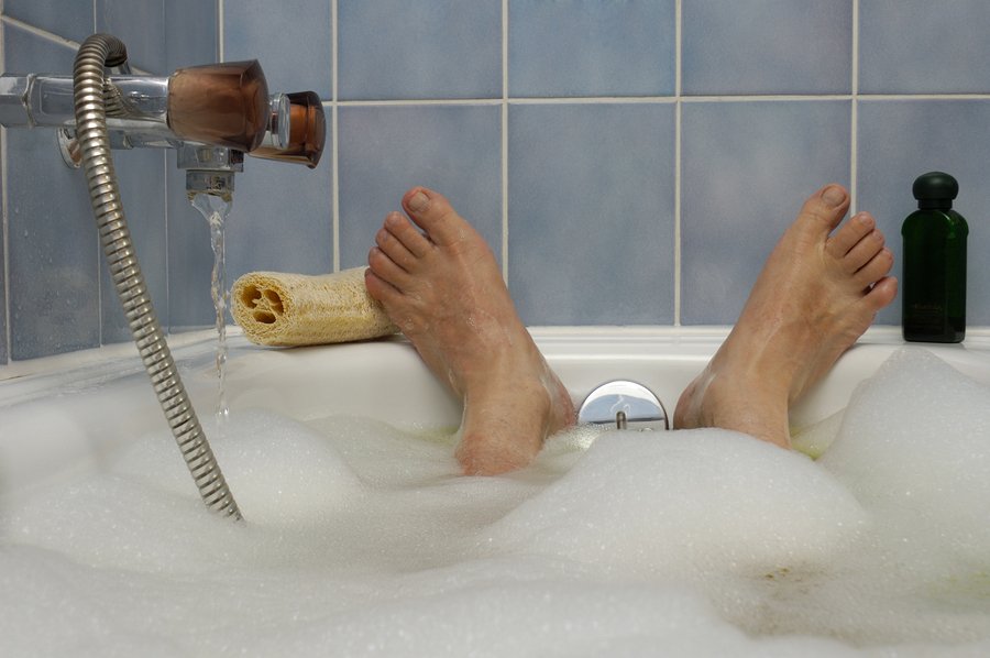 A man's feet, poking out of a bubble bath, with water running in to the side. A loofah and bottle of bath lotion to either side. Space for text on the blue tiled wall above the feet.